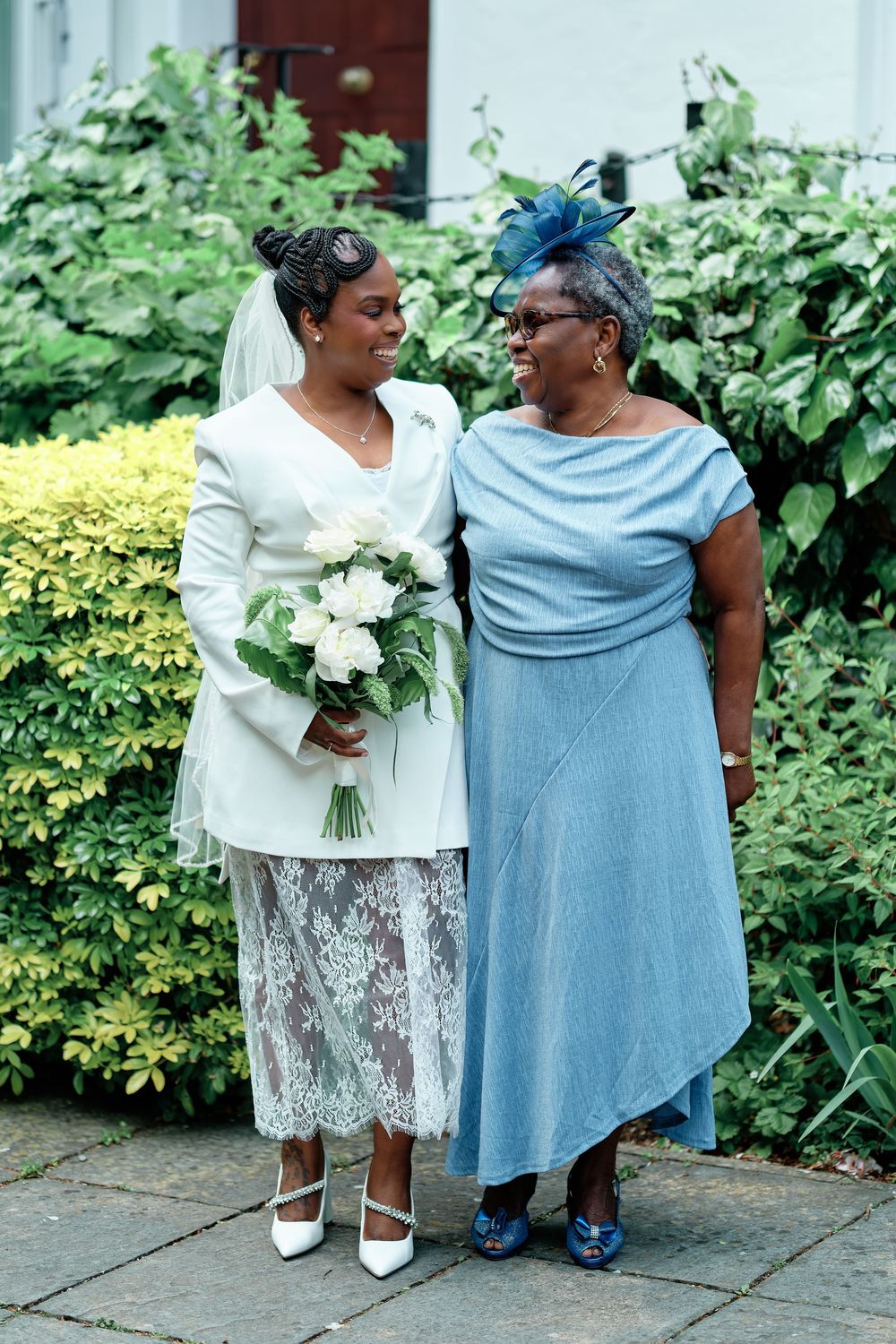 Bride in white lace dress holding bouquet stands with older woman in blue dress and fascinator outdoors.