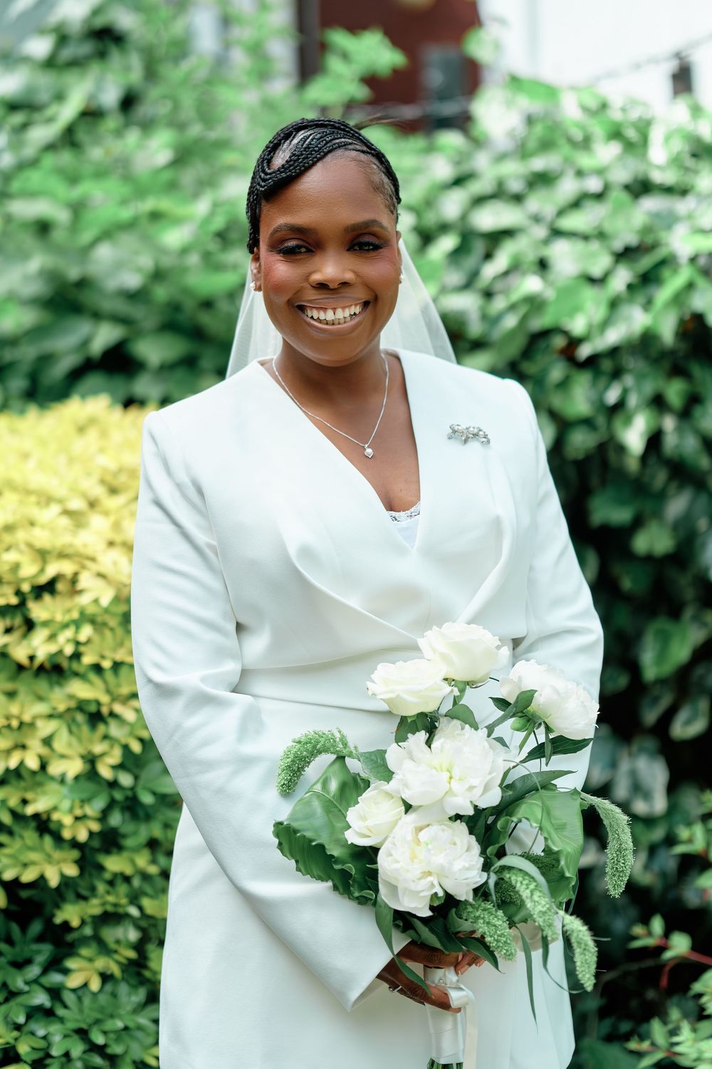 Smiling Black bride in white blazer dress holding white floral bouquet, wearing veil outdoors with green foliage background.