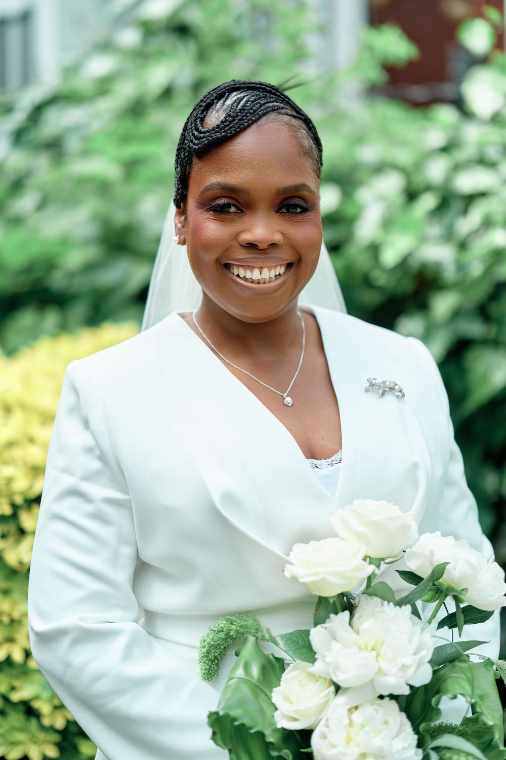 Smiling bride in white suit holding white bouquet with veil and braided hair outdoors.