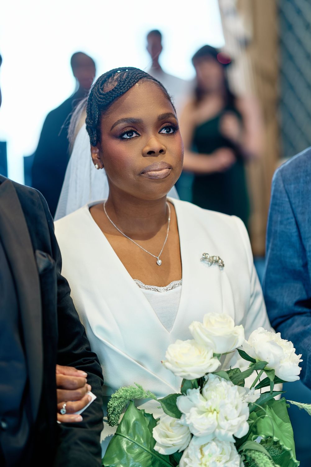 Elegant Black bride in white blazer holding white peonies, wearing veil and brooch at wedding ceremony.