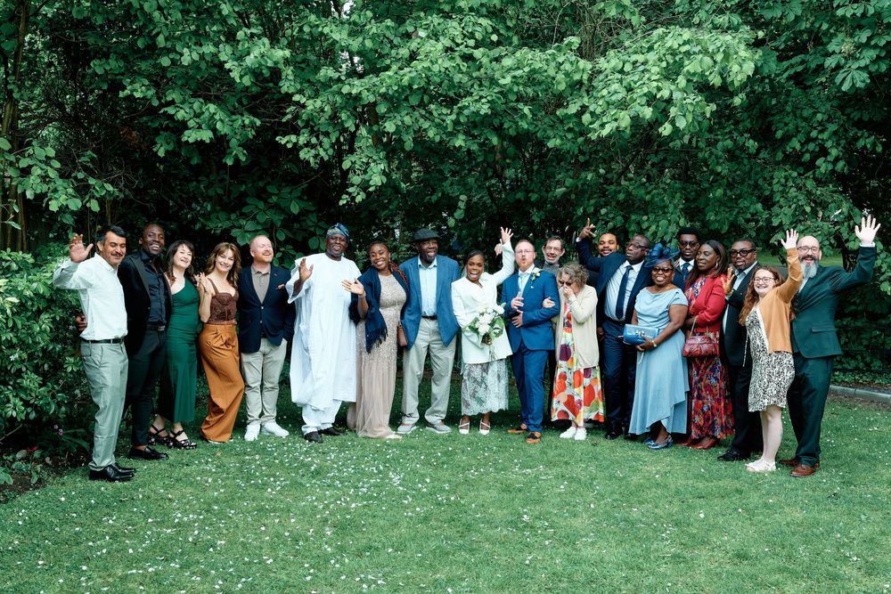 Large diverse group of people posing together outdoors in a lush green garden, waving and smiling at a wedding celebration.