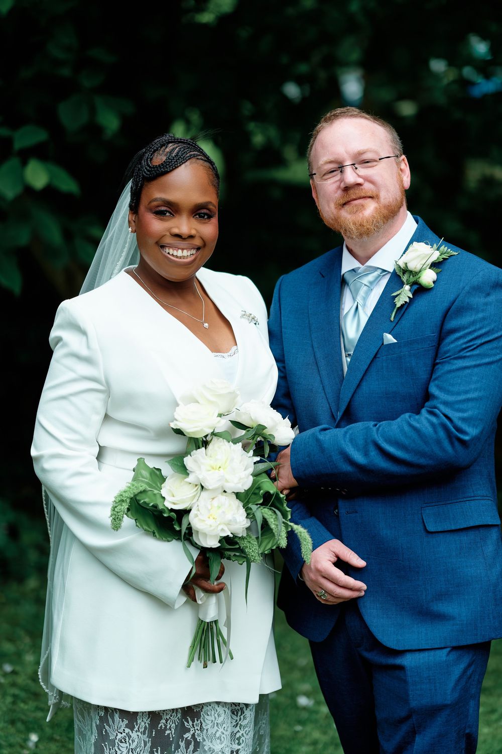 Bride in white blazer holding bouquet stands with groom in blue suit outdoors at wedding ceremony.