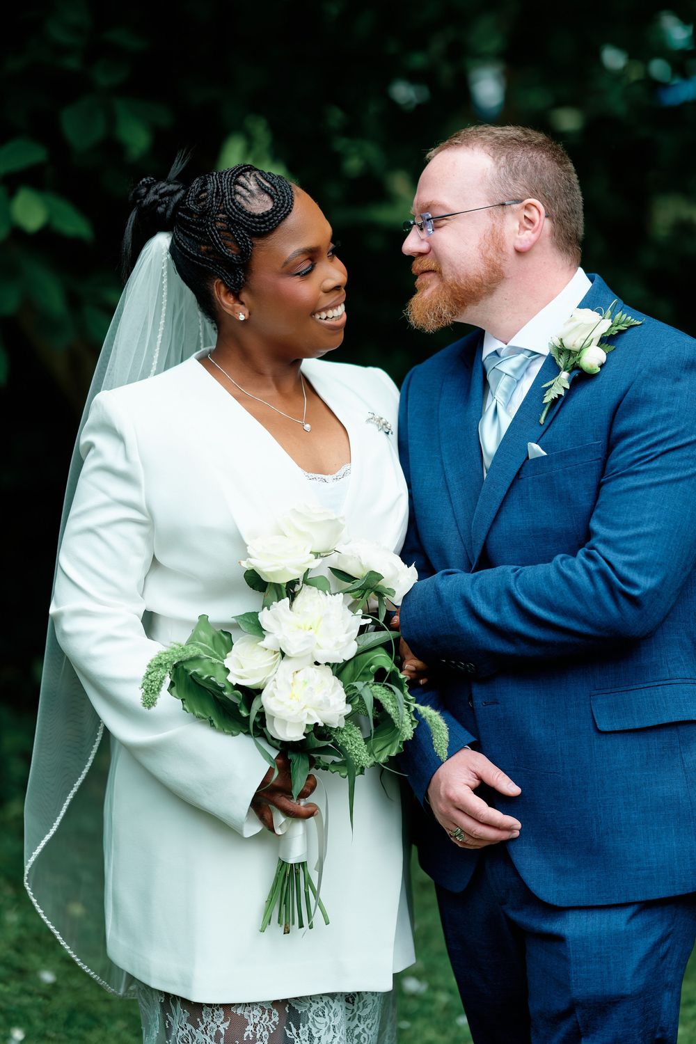 A bride and groom share a joyful moment outdoors, she holding white peonies in a white blazer, he in a navy suit.