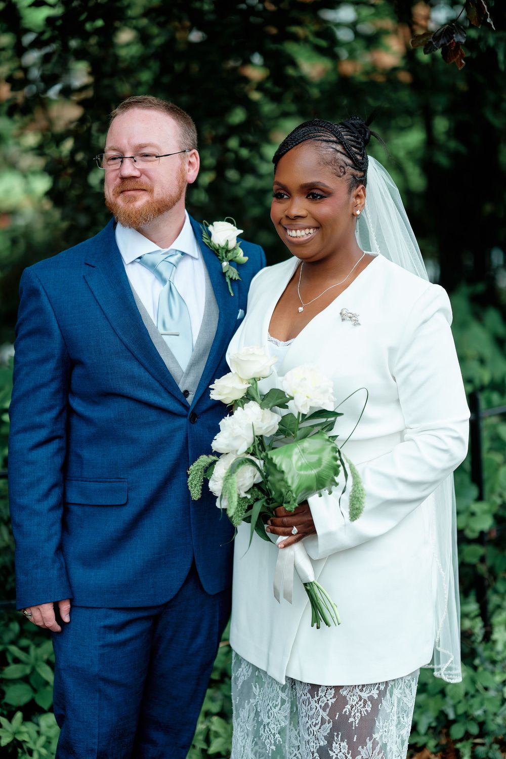 Interracial couple posing for wedding portrait, groom in blue suit, bride in white blazer holding white rose bouquet.