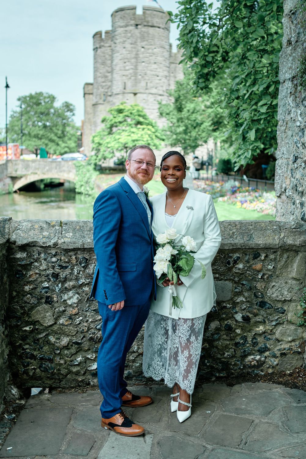 Couple in wedding attire pose by stone wall with river and historic tower backdrop on their wedding day.
