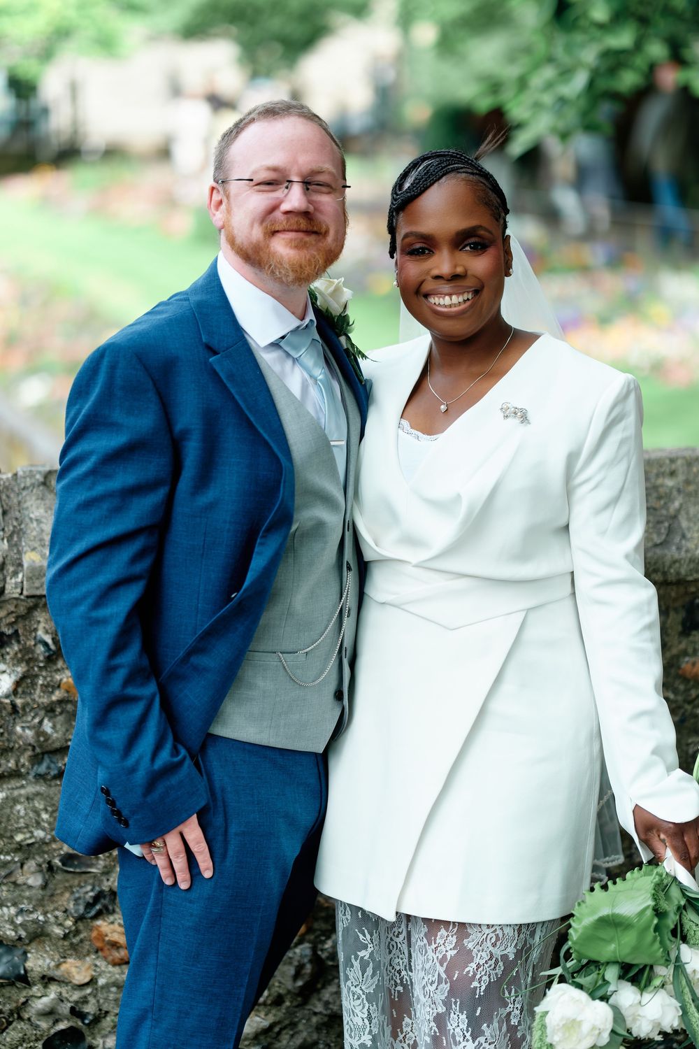 A smiling couple in wedding attire, man in blue suit and woman in white jacket with lace skirt, posing outdoors.
