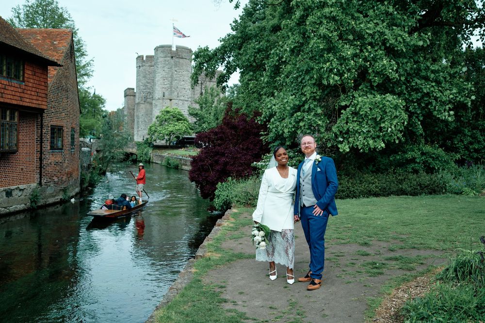 Newlywed couple posing by a scenic river with a historic castle and punting boat in the background.