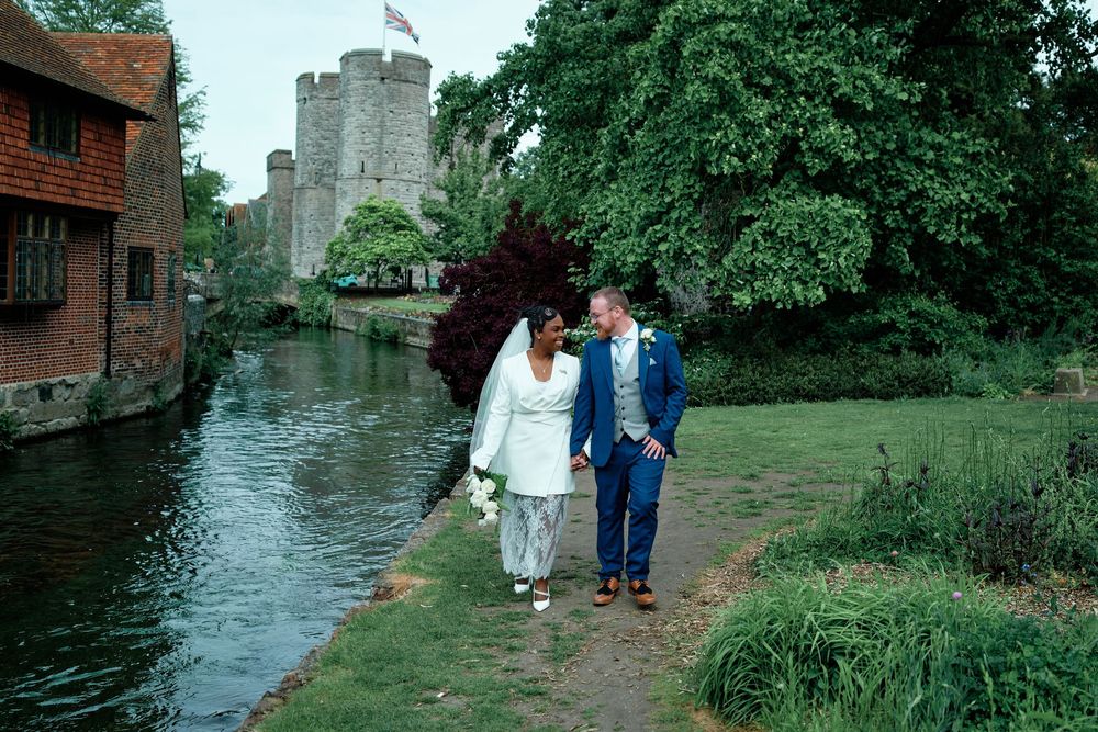 Bride and groom walking along river path by historic castle on wedding day.