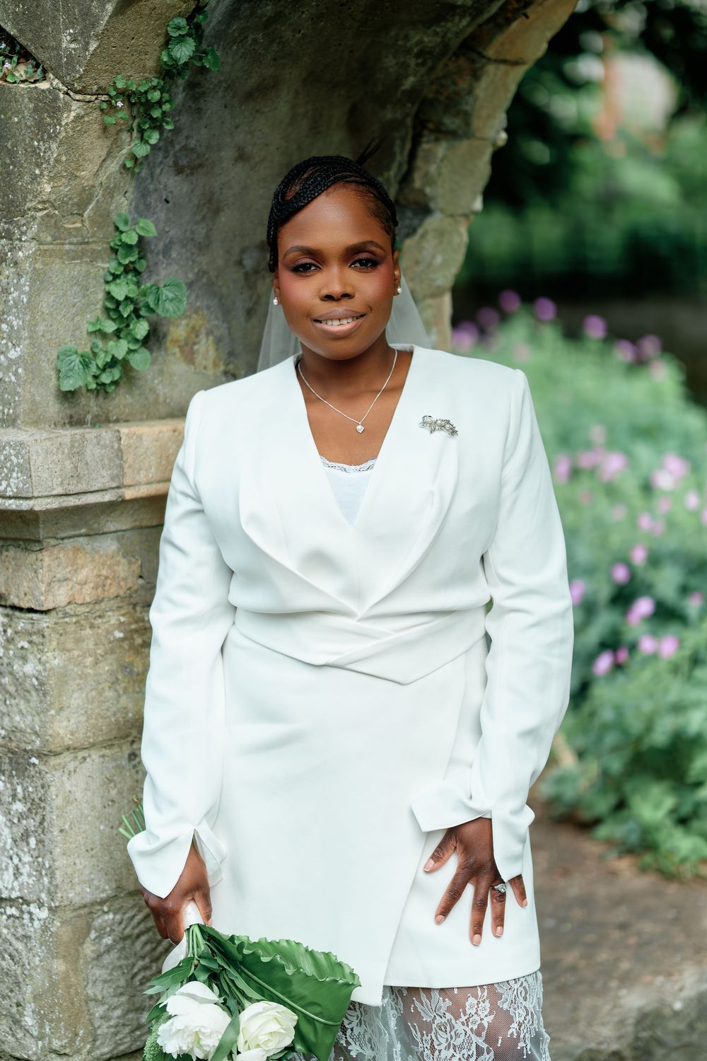 Black bride in white blazer dress with brooch holds white bouquet by stone arch with ivy.