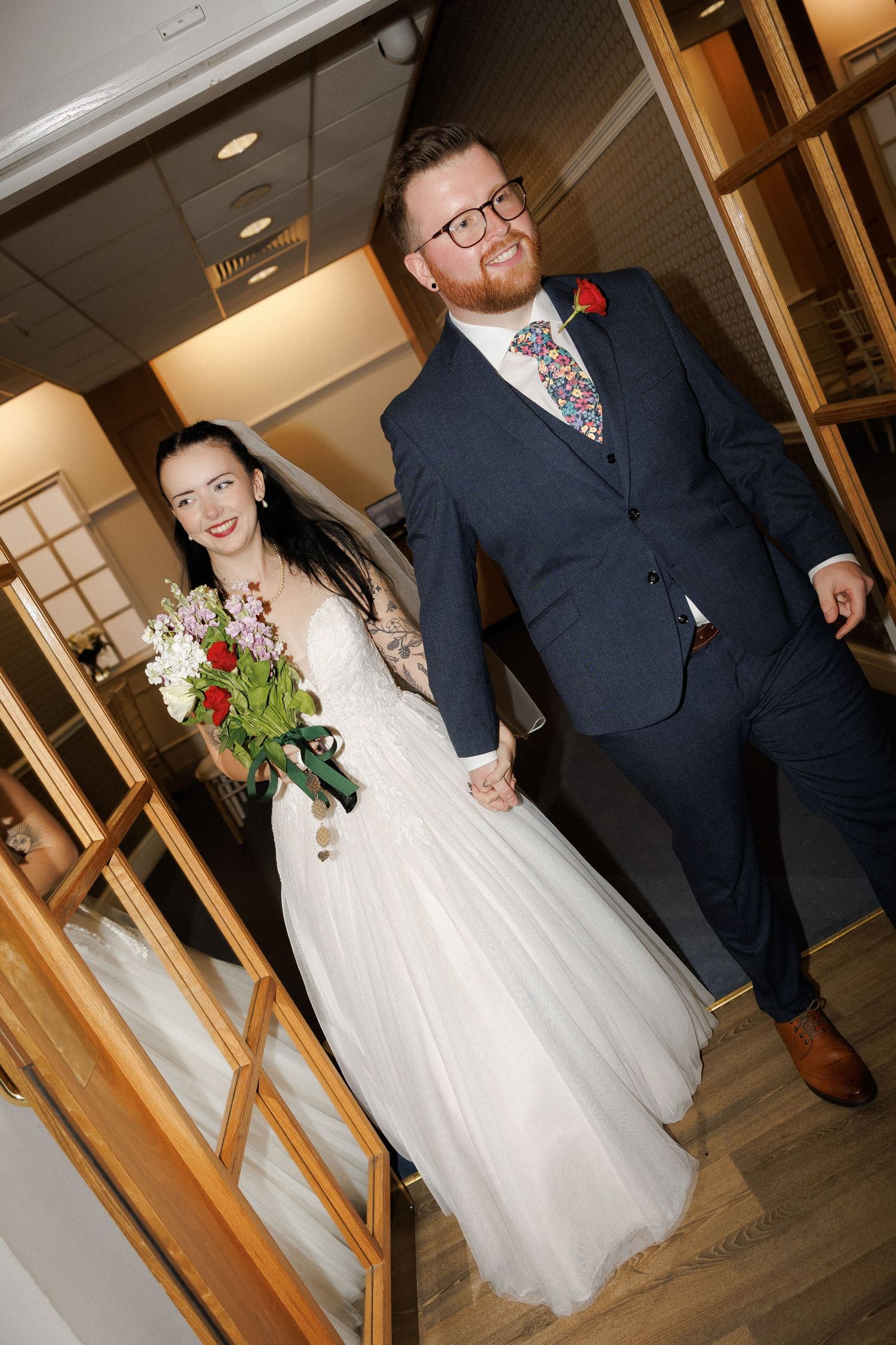 A smiling bride in a white gown holding a bouquet walks hand-in-hand with a bearded groom in a navy suit.