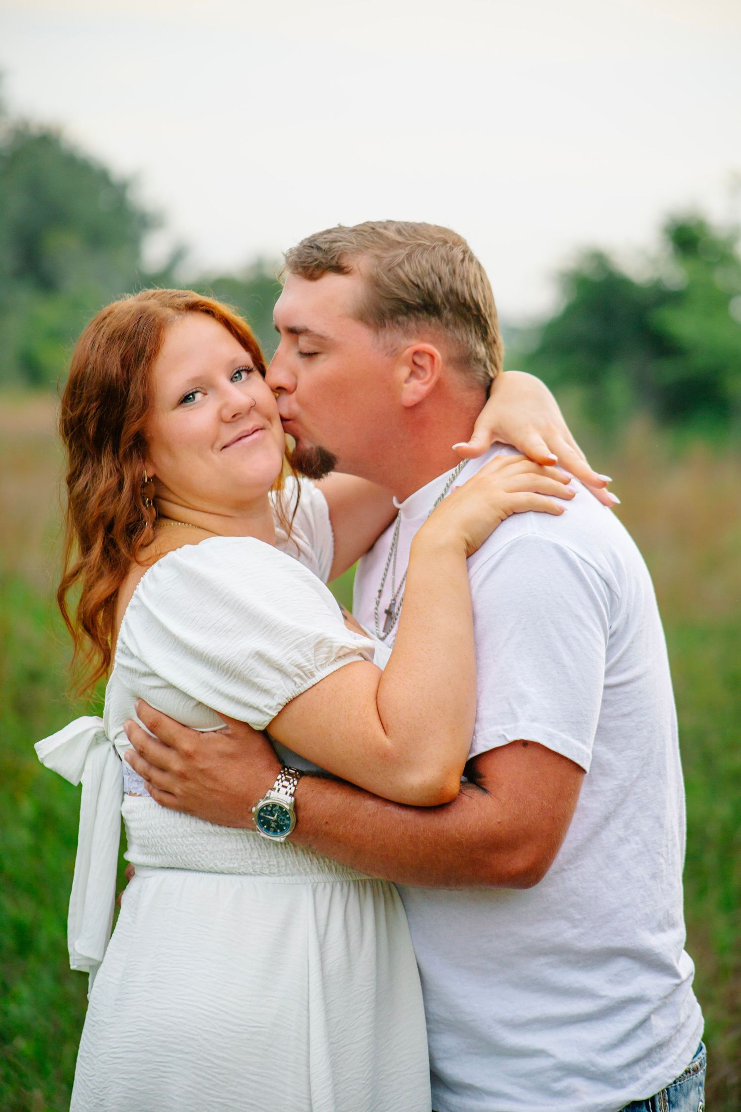 A man tenderly cups a smiling redhead woman's face in an outdoor field setting.