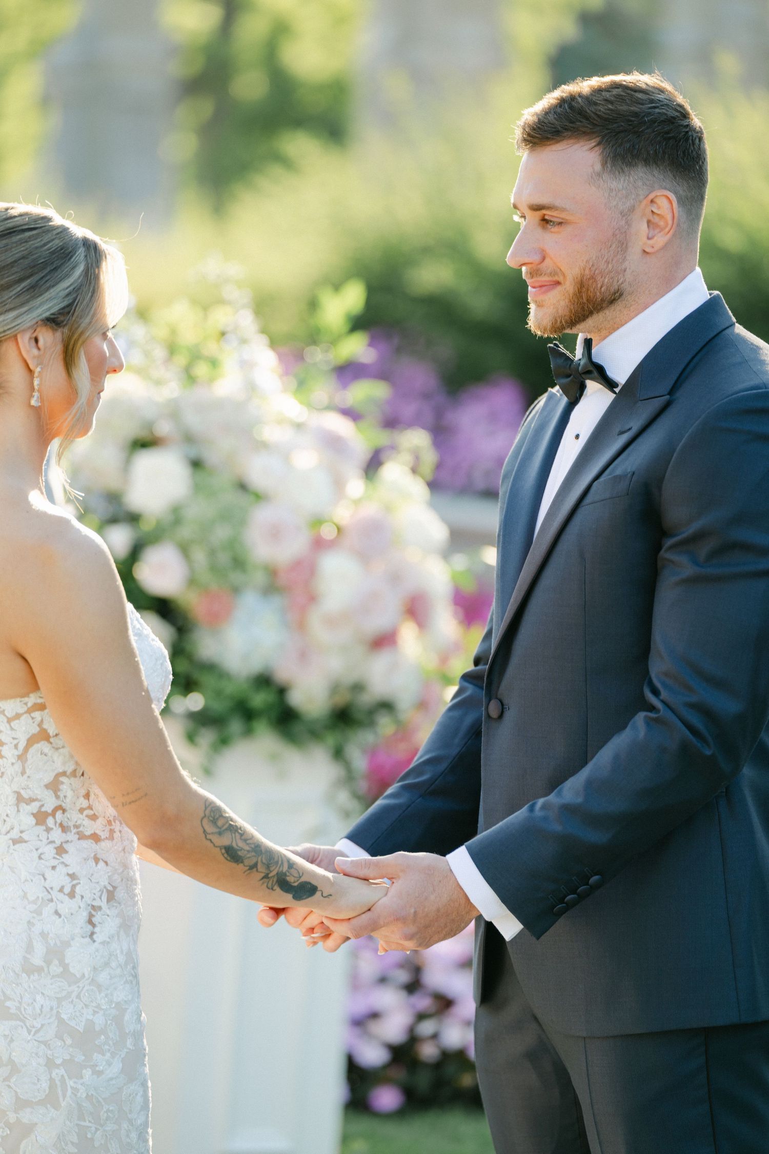 Armour House Wedding - Groom in navy tuxedo holds bride's hands during outdoor wedding ceremony with floral arch backdrop.