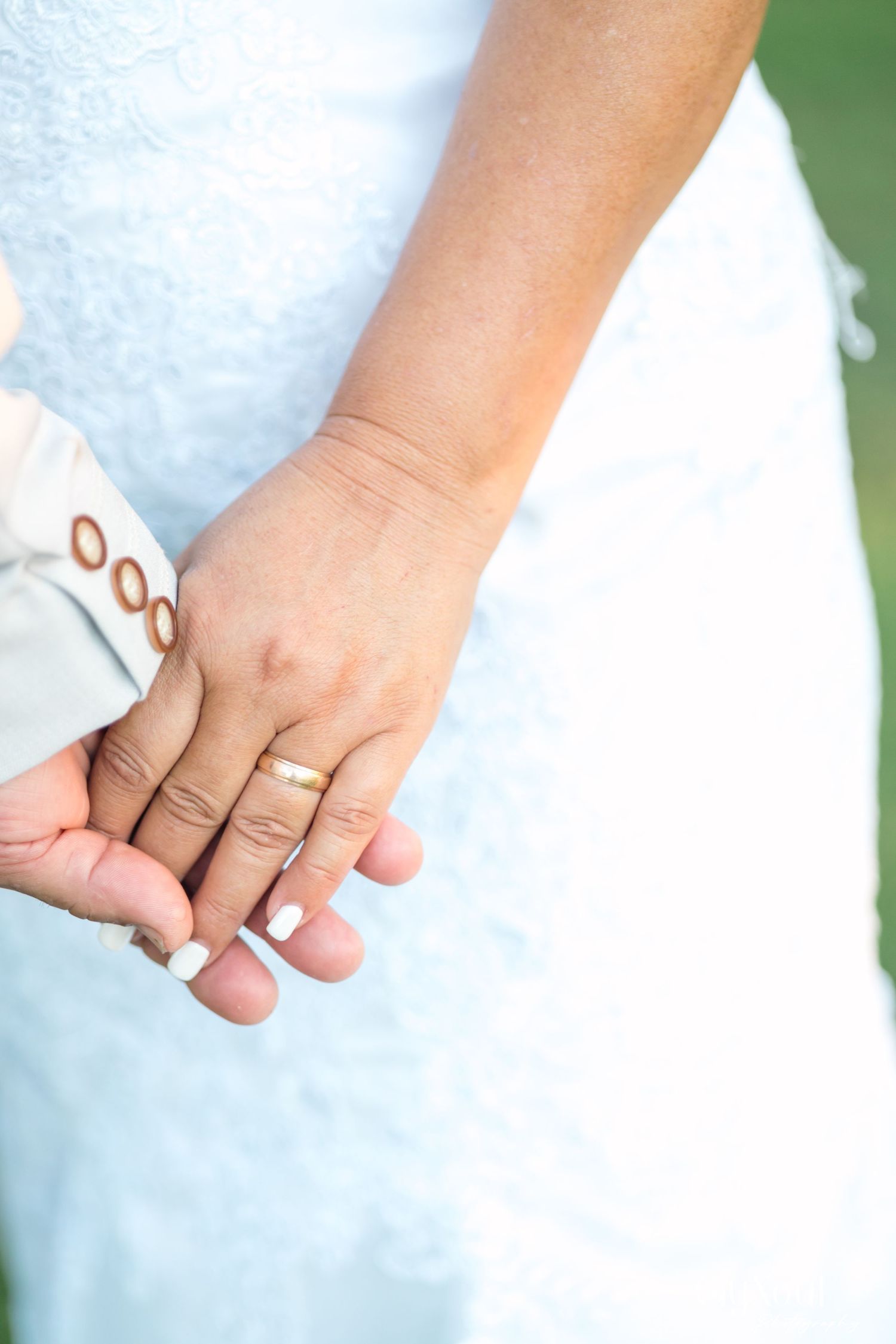 Close-up of newlywed couple holding hands, bride wearing wedding ring and white lace dress.