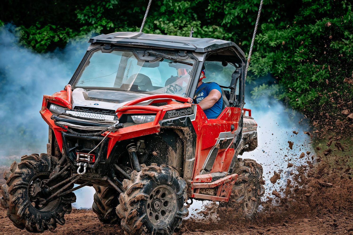 Red UTV side-by-side off-road vehicle splashing mud on a dirt track with a driver in blue helmet.