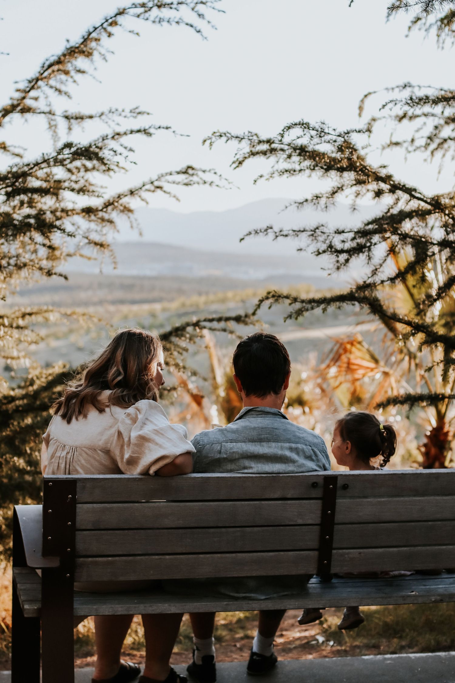 Family of three sitting on a bench enjoying a scenic mountain valley view framed by acacia trees at sunset.