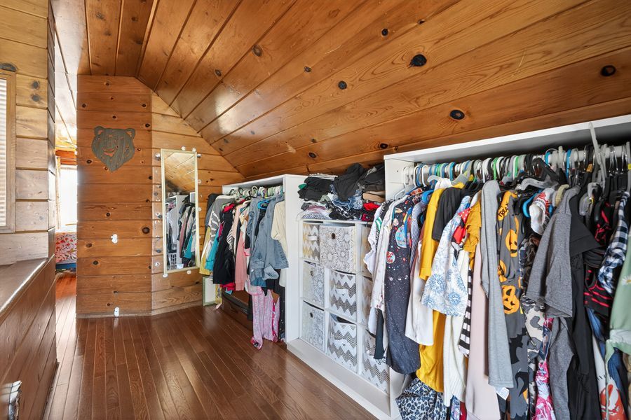 Spacious walk-in closet with wood-paneled walls and ceiling, featuring double hanging rods filled with clothing and hardwood floors.