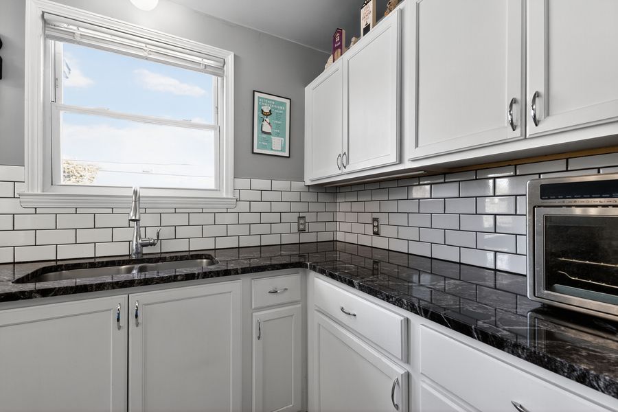 White kitchen with subway tile backsplash, dark granite countertops, and stainless steel appliances near a bright window.