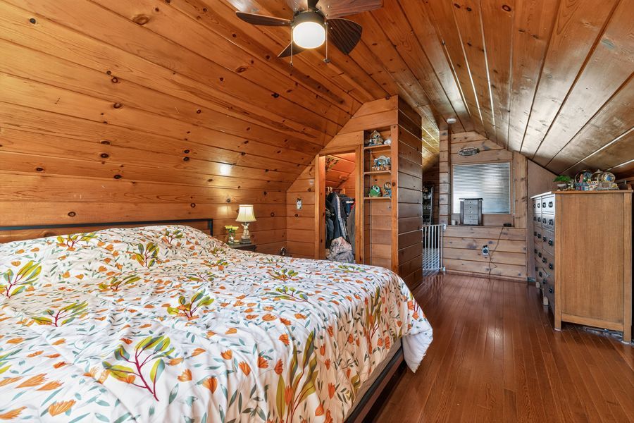 Cozy loft bedroom with wood-paneled walls, vaulted ceiling, hardwood floors, and a floral patterned bedspread.