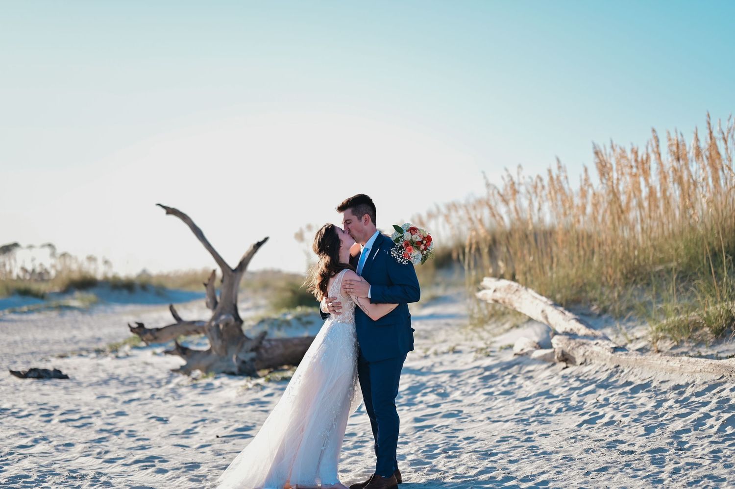 Bride and groom share a romantic embrace on a sandy beach with sea oats and driftwood in the background.