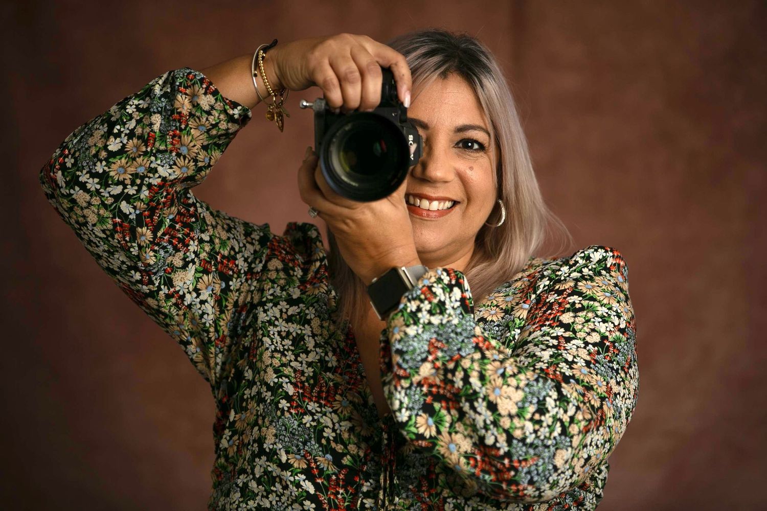 Woman in floral blouse smiling while holding a DSLR camera up to her eye in a studio setting.