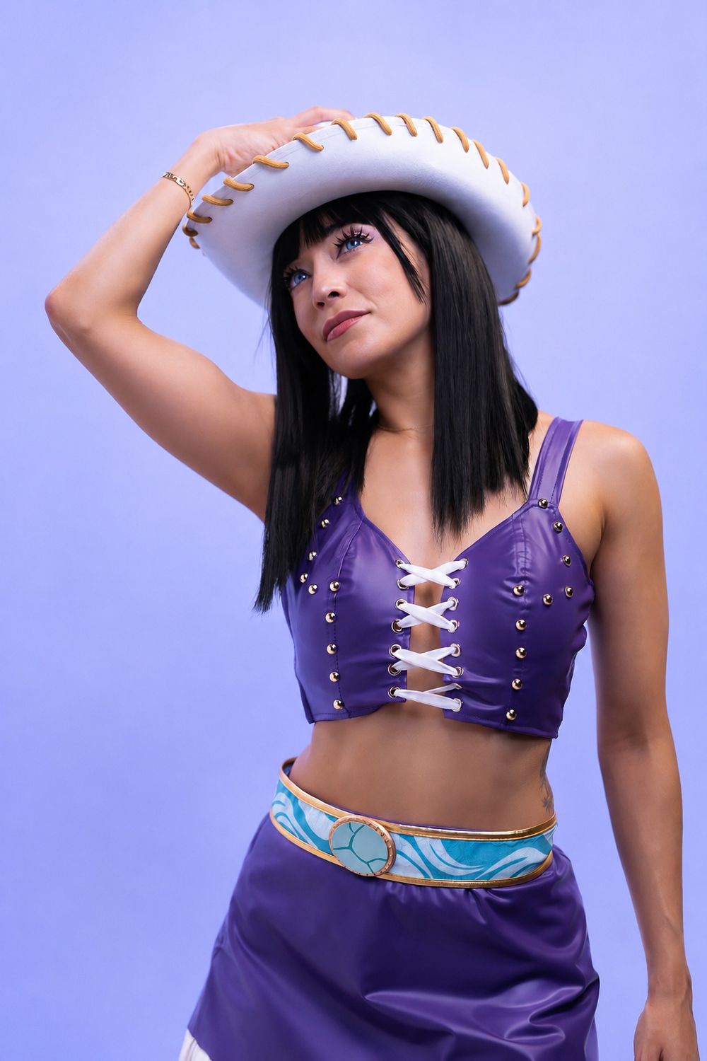 Woman in purple crop top and skirt with white cowboy hat poses against lavender background.