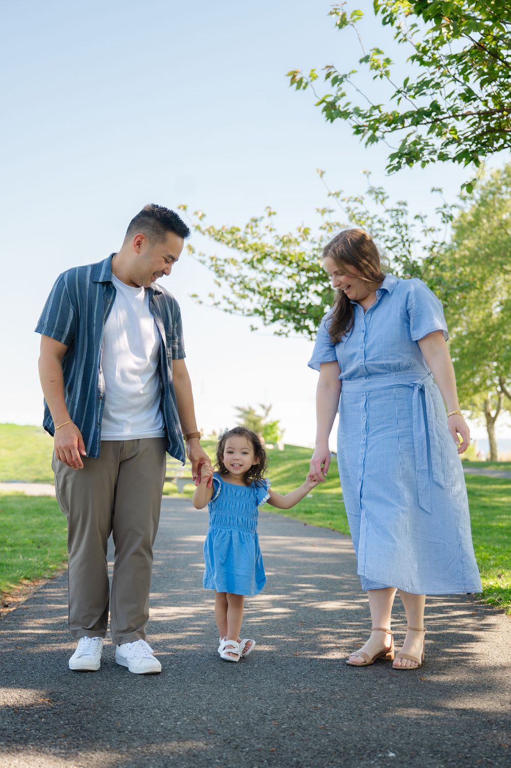 A family of three walks hand-in-hand in a sunny park, parents smiling at their young daughter in a blue dress.