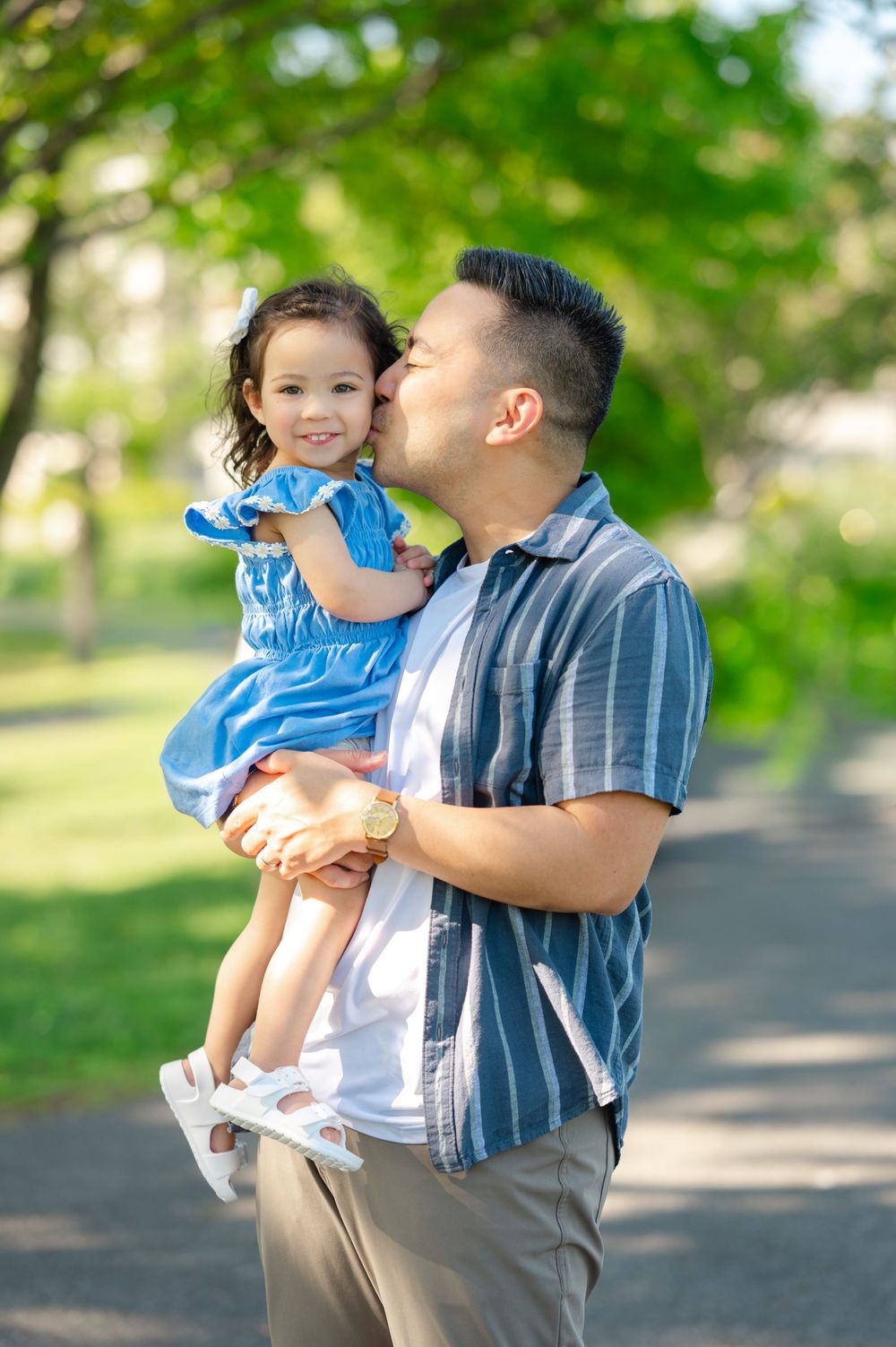 A loving father kisses his happy toddler daughter in a blue dress in a sunny green park.