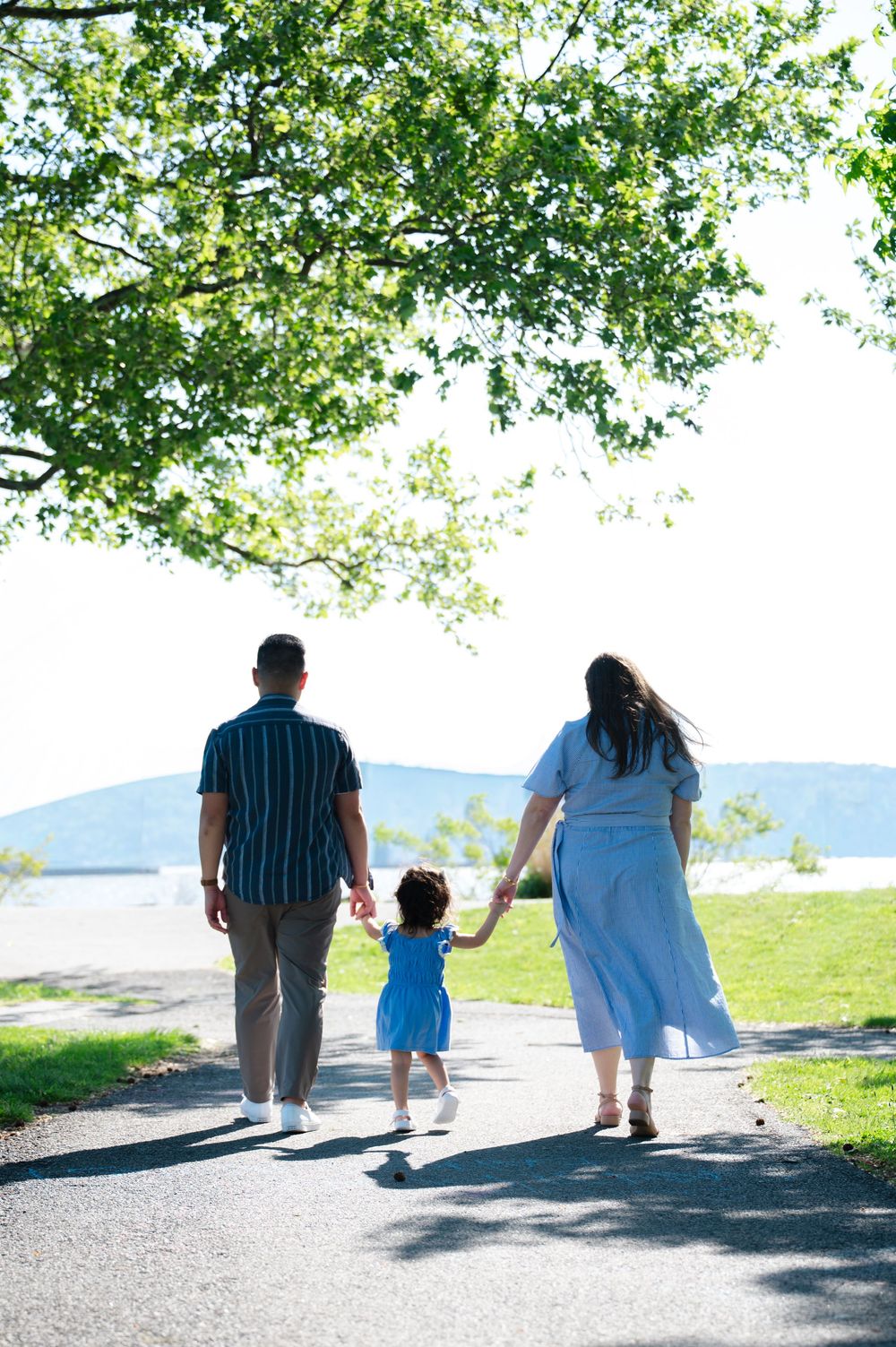 A family of three walks hand-in-hand along a sunny park path near a scenic waterfront.