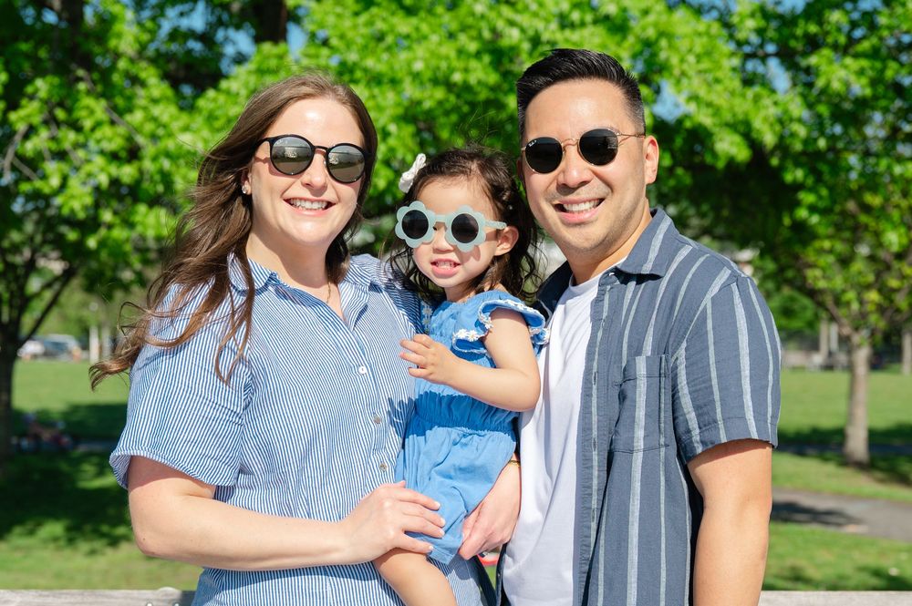 Happy family of three wearing sunglasses smiling outdoors in a sunny park with lush green trees.