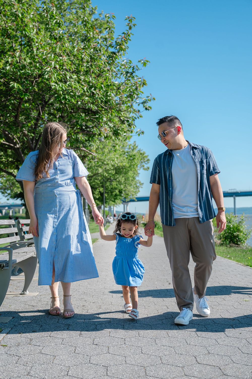 Family of three walking hand-in-hand on sunny park path, parents holding toddler girl in blue dress between them.