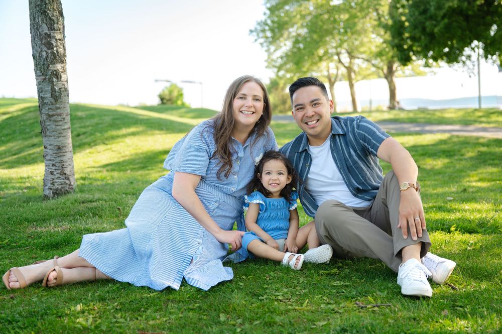 Happy family of three sitting on green grass in a sunny park, parents smiling with young toddler daughter between them.