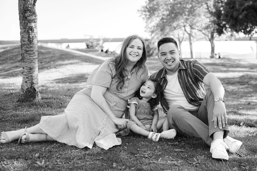 Black and white photo of a happy family with parents and young daughter sitting on grass in a park.