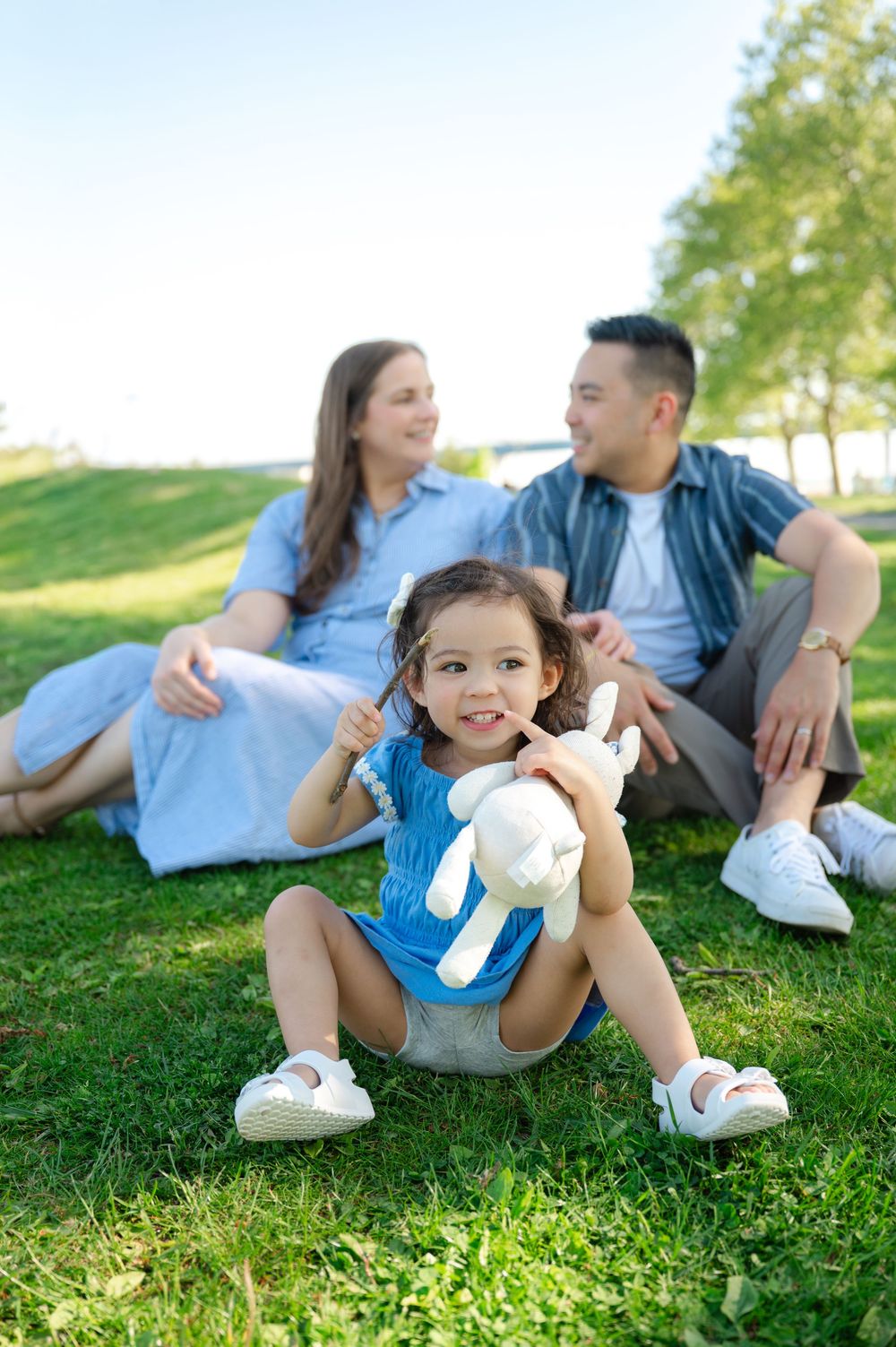 A happy family sits on green grass in a park, with their toddler girl holding a stuffed bunny toy.
