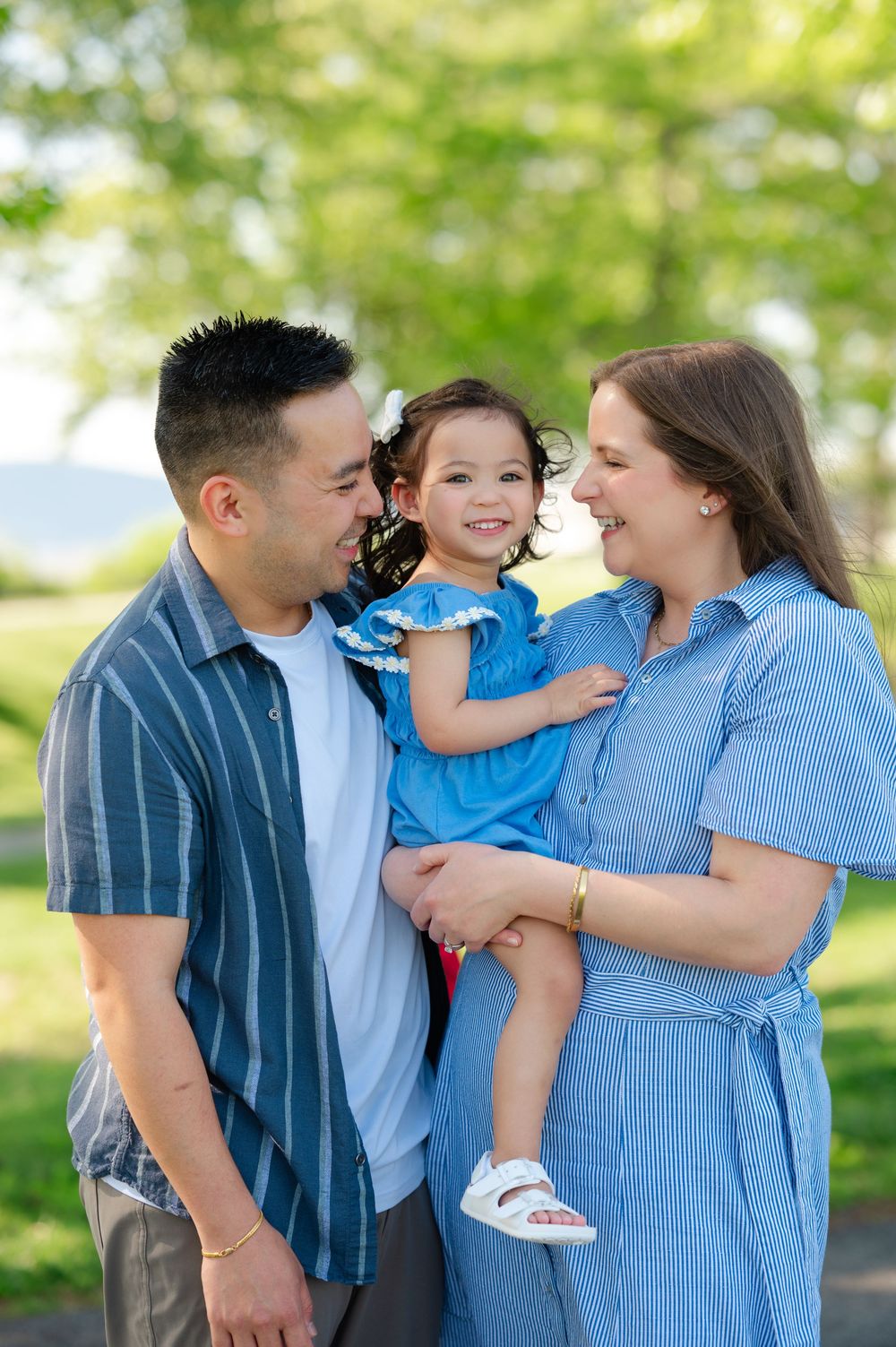 A happy family of three poses outdoors, parents smiling at their young daughter in a blue dress amid green trees.