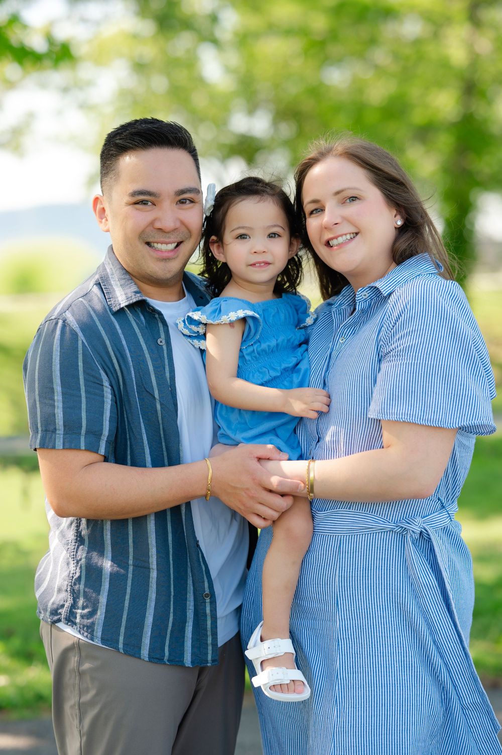 A smiling family of three poses outdoors, parents in blue outfits holding their young daughter in a green park setting.