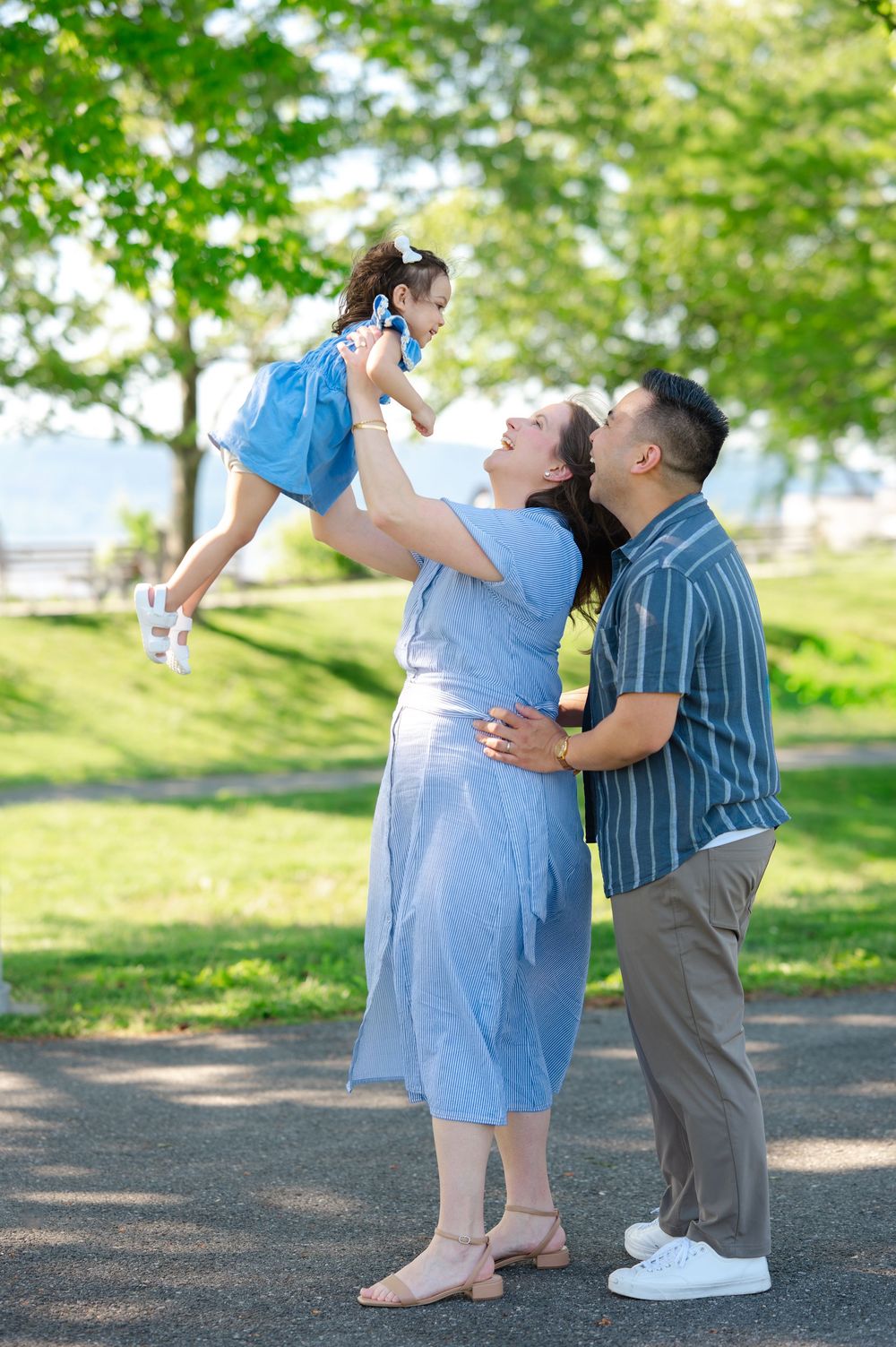 A joyful family in a park, mother lifting daughter in blue dress while father embraces them both.