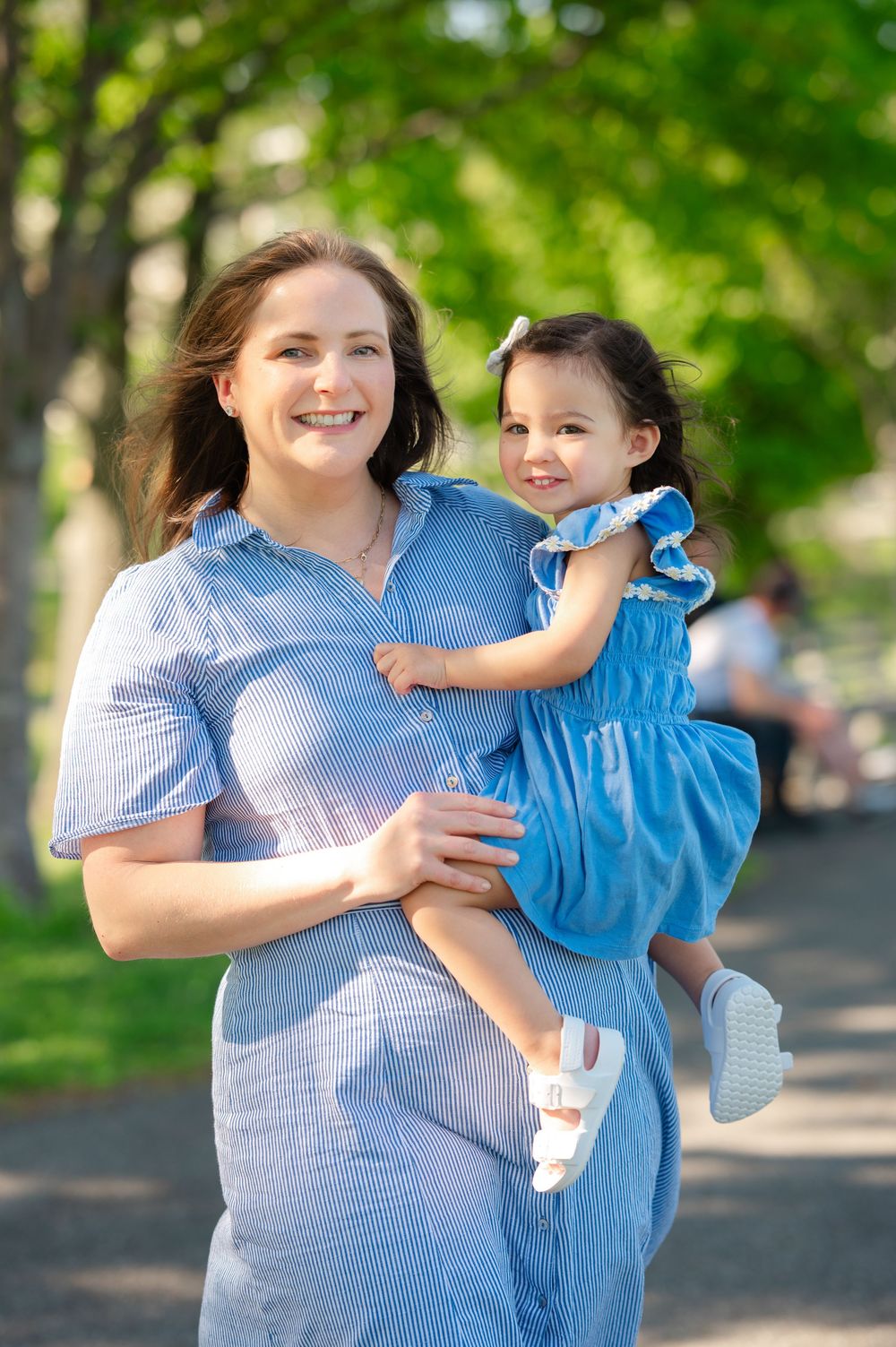 A smiling mother in a blue dress holds her toddler daughter in a matching blue dress at a sunny park.