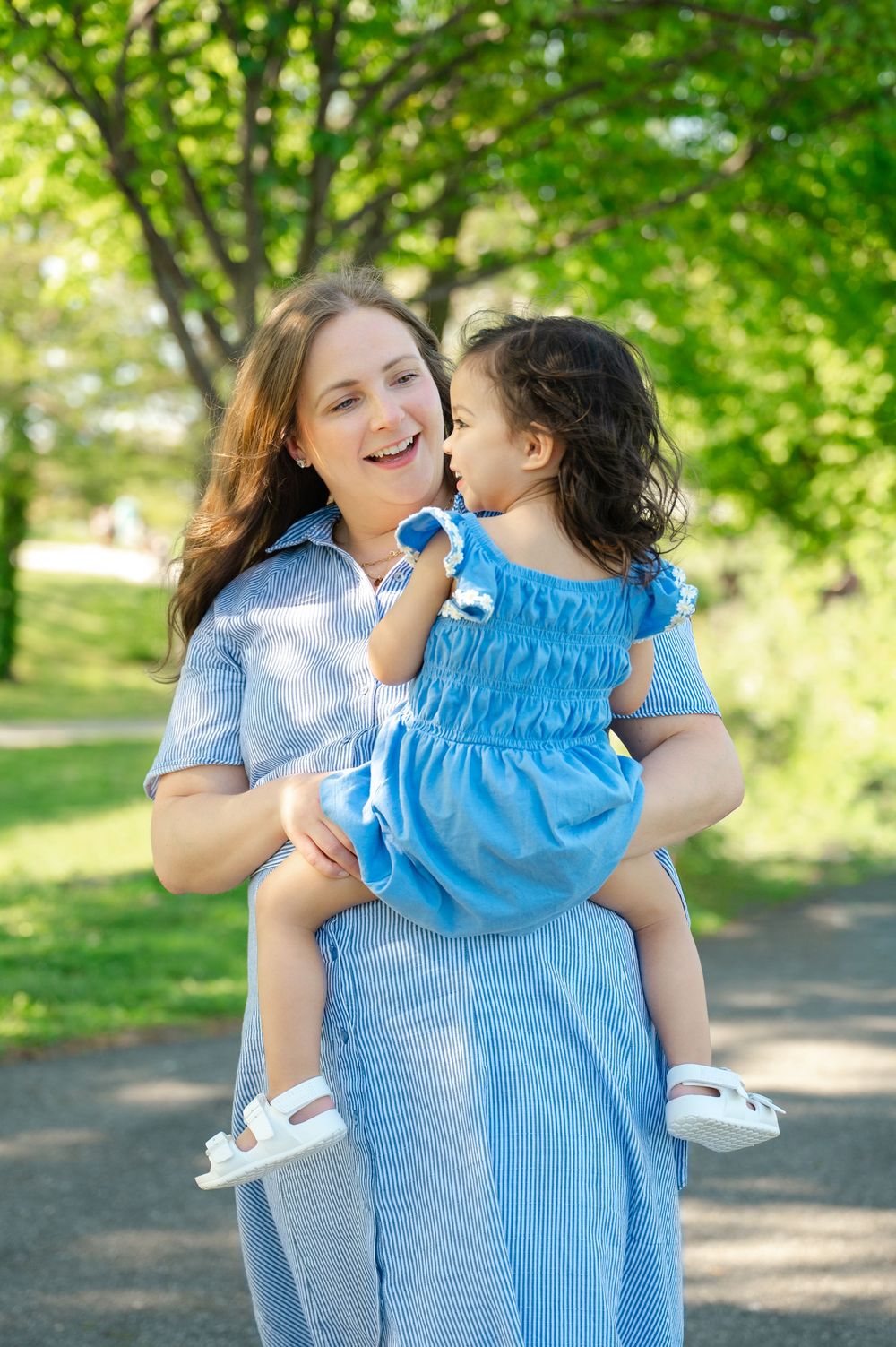 A smiling mother in a blue dress holds her toddler daughter in a matching blue outfit in a sunny park.