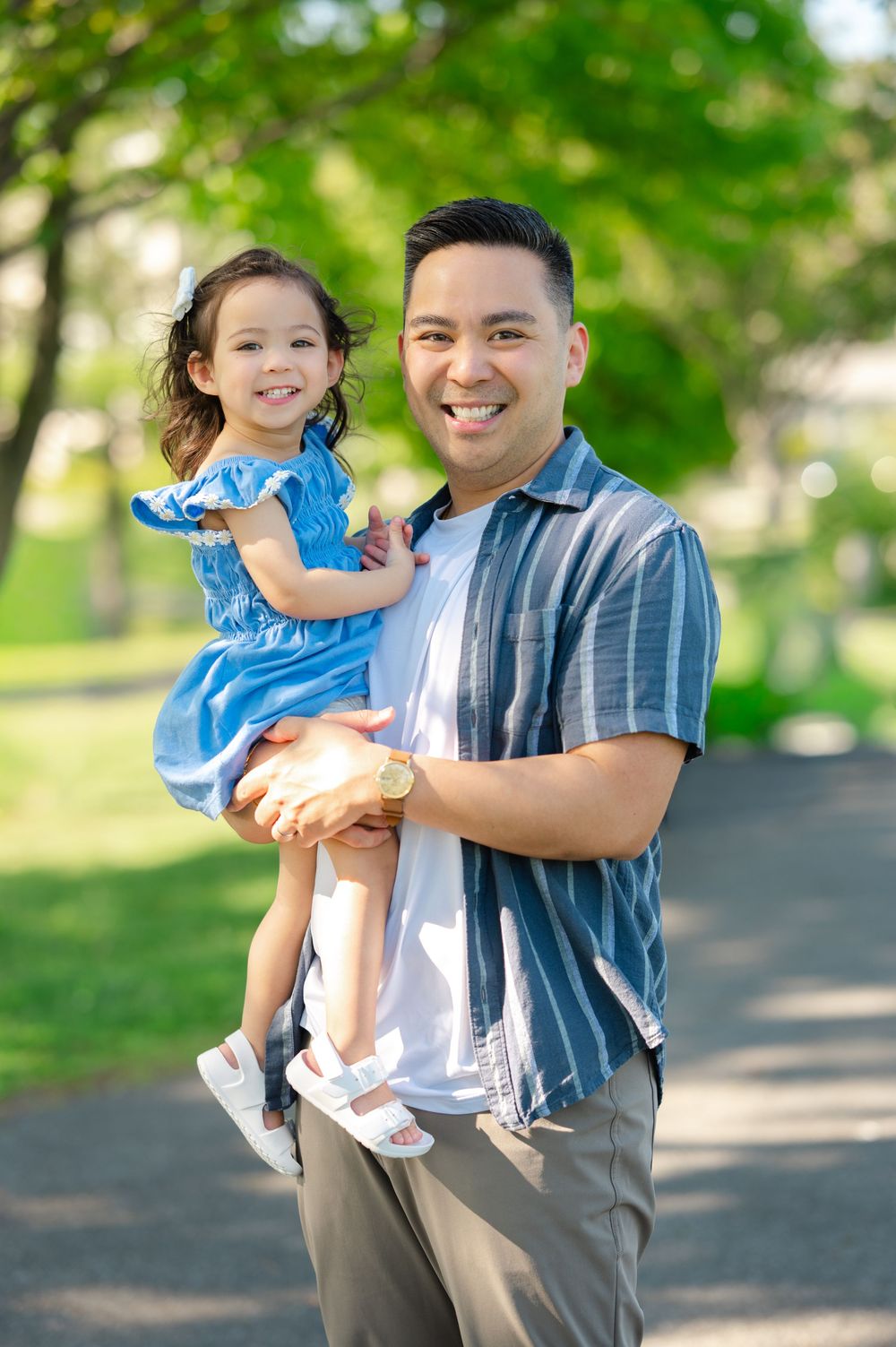 A smiling man holds a young girl in a blue dress outdoors in a sunny green park.