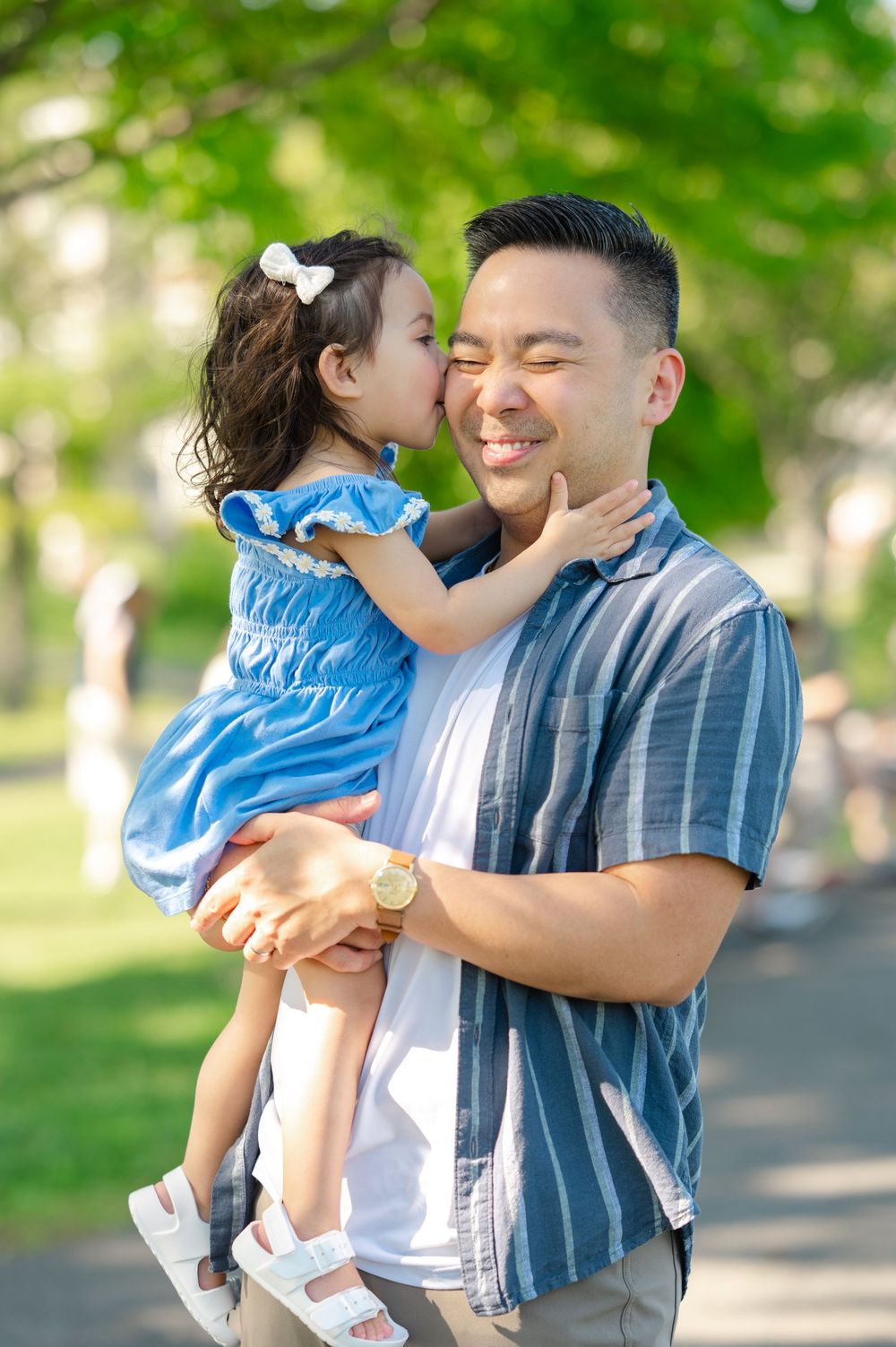 A young girl in a blue dress gives her smiling father a sweet kiss on the cheek outdoors.
