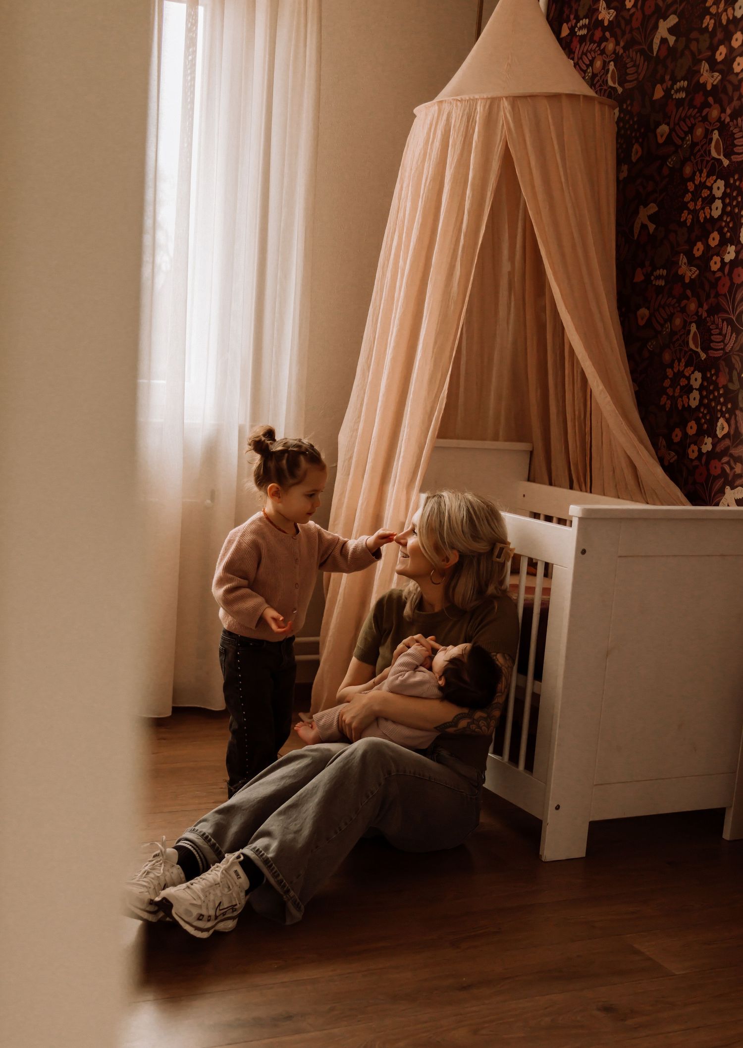 A man leans against a wall watching two women holding babies in a cozy nursery with warm wood floors.