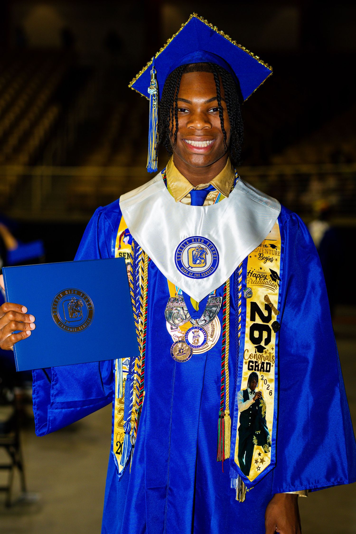 Smiling graduate in blue cap and gown holds diploma, wearing medals and decorated stole at graduation ceremony.
