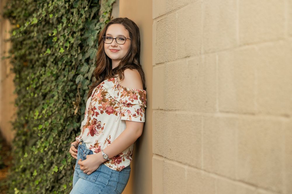 Young woman in floral off-shoulder top and jeans poses against ivy-covered stone wall outdoors.