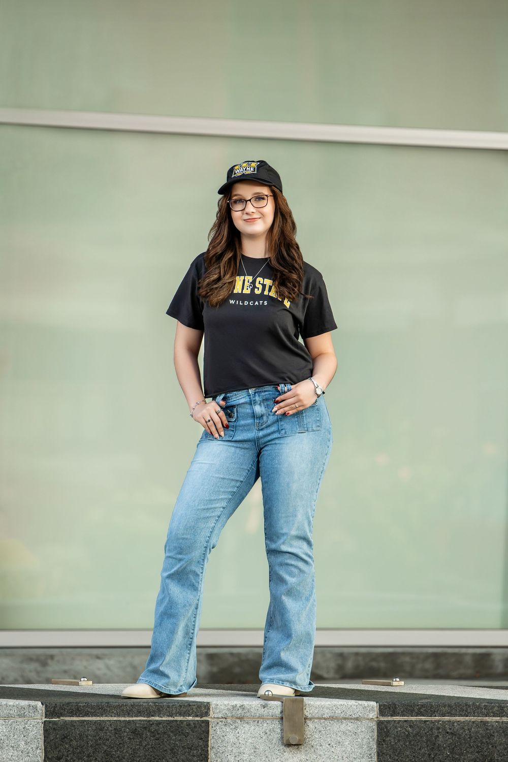 Young woman in Wayne State Wildcats shirt, jeans, and cap posing outdoors near a glass wall.