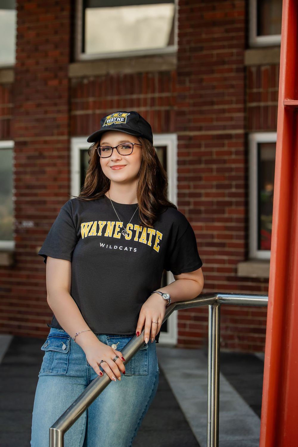 Young woman in Wayne State Wildcats shirt and cap leaning on a railing outside a brick building.