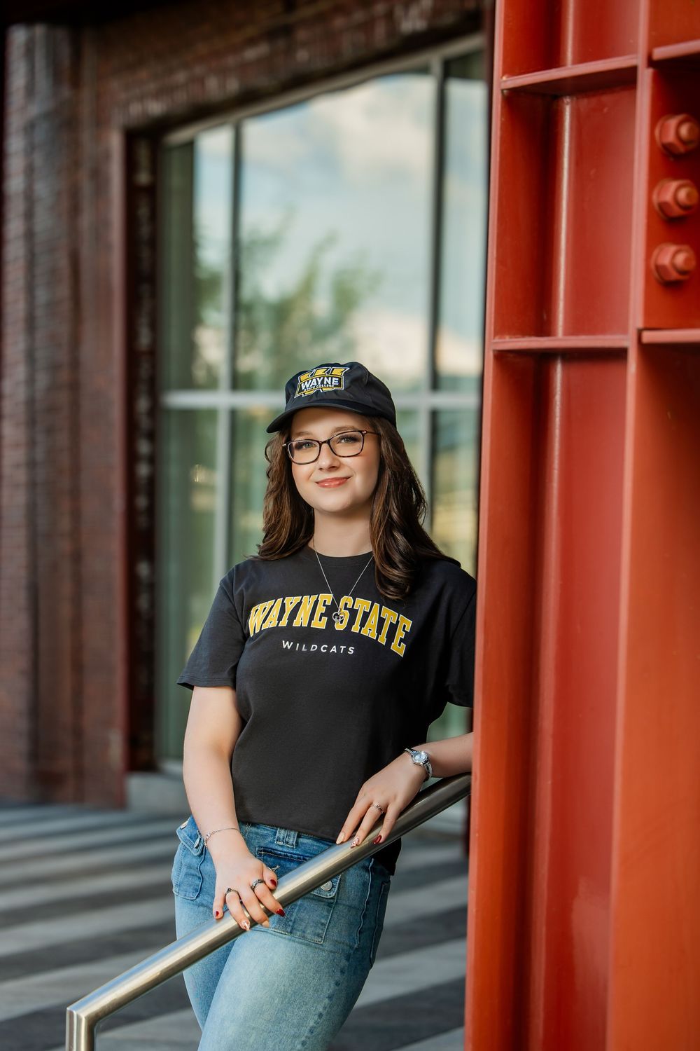 Young woman in Wayne State Wildcats shirt and cap leans on railing near red steel structure.