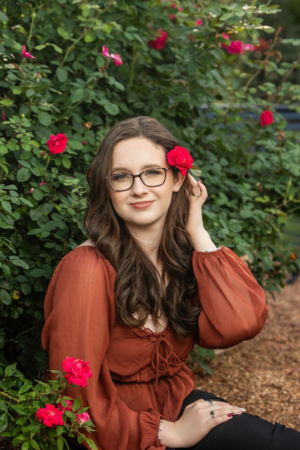 Young woman in rust blouse holds red rose near blooming rose garden bushes outdoors.