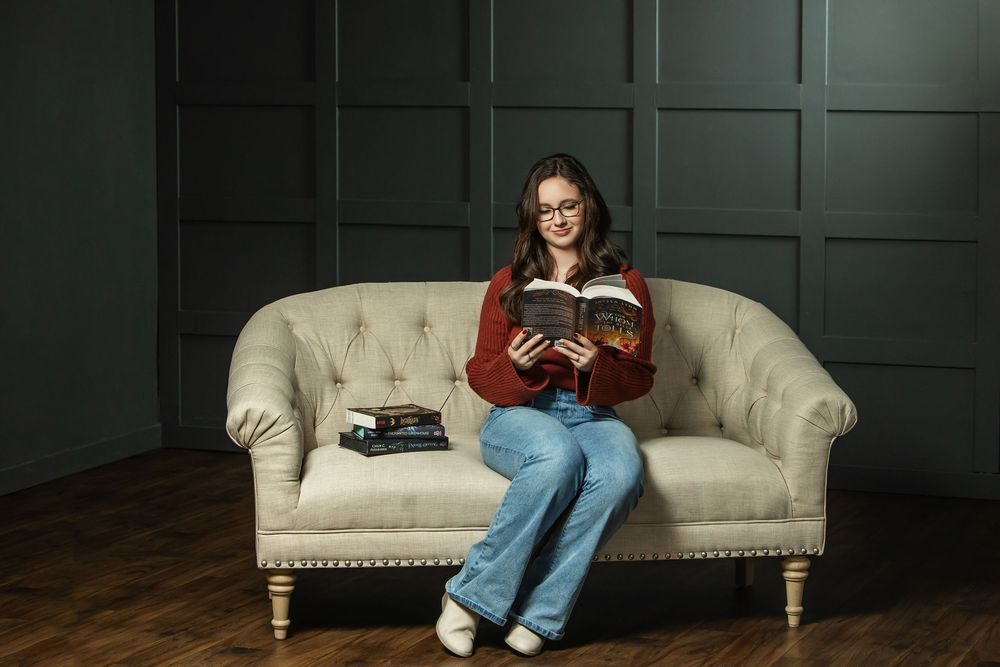 Young woman with glasses reading a book on a beige tufted sofa in a dark-walled studio.