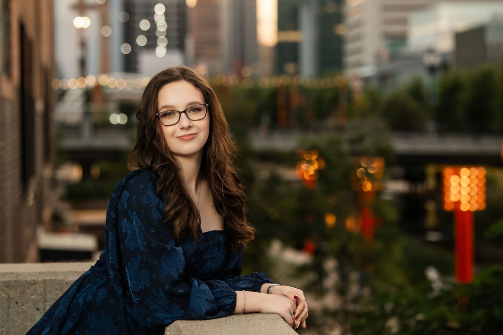 Young woman with glasses and dark hair posing at dusk in a blue dress with city lights behind her.