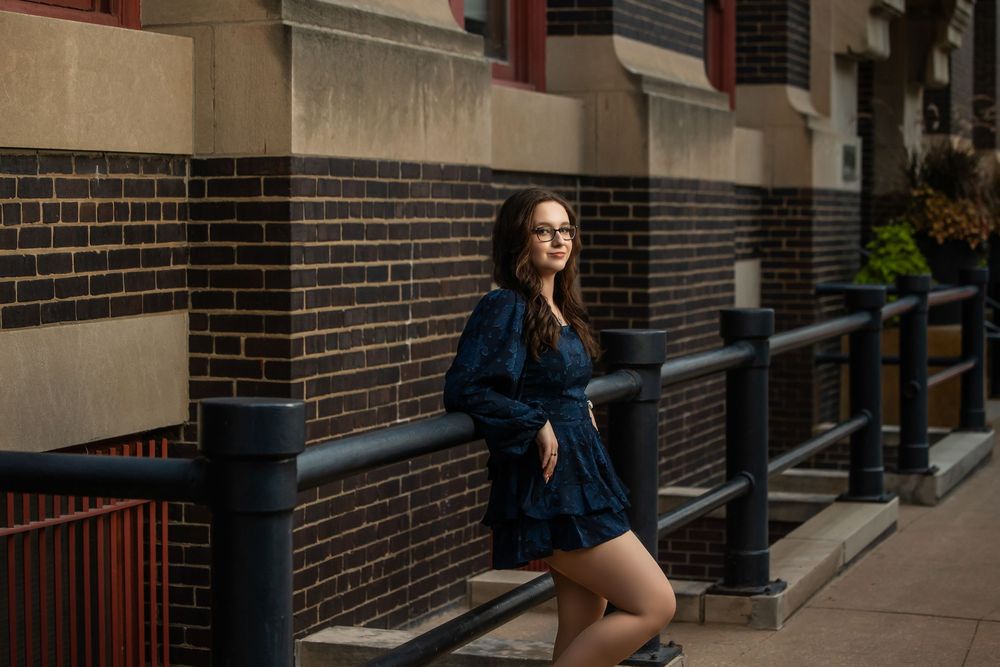 Young woman in navy blue romper leans on black railing outside brick building.